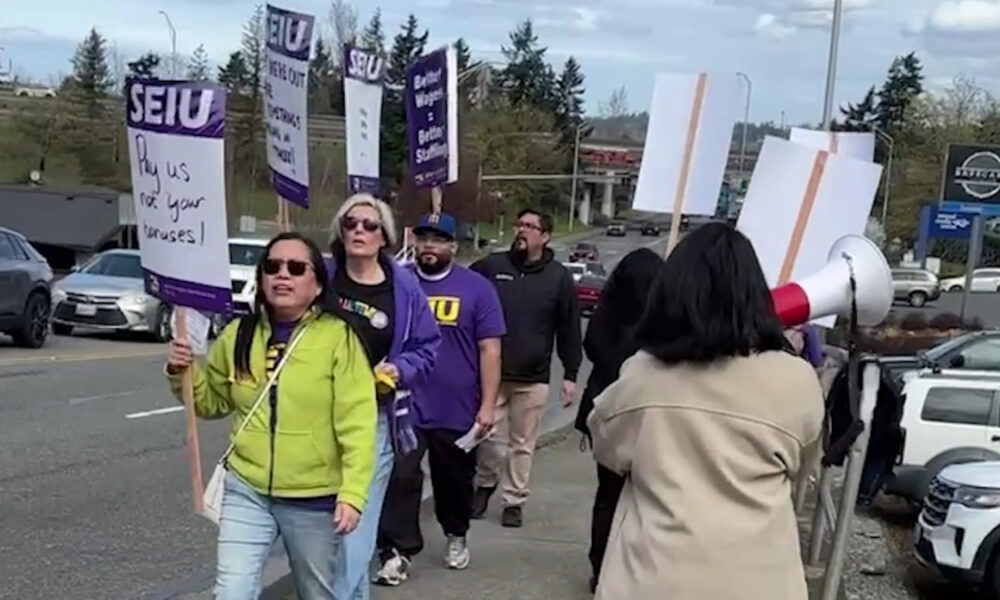 Group of protesters marching on a street, holding SEIU signs; a woman in a lime jacket leads with a sign. Meets a megaphone on the right.