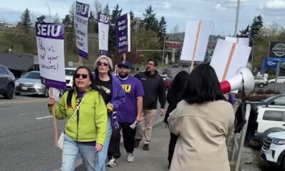 Group of protesters marching on a street, holding SEIU signs; a woman in a lime jacket leads with a sign. Meets a megaphone on the right.