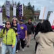 Group of protesters marching on a street, holding SEIU signs; a woman in a lime jacket leads with a sign. Meets a megaphone on the right.