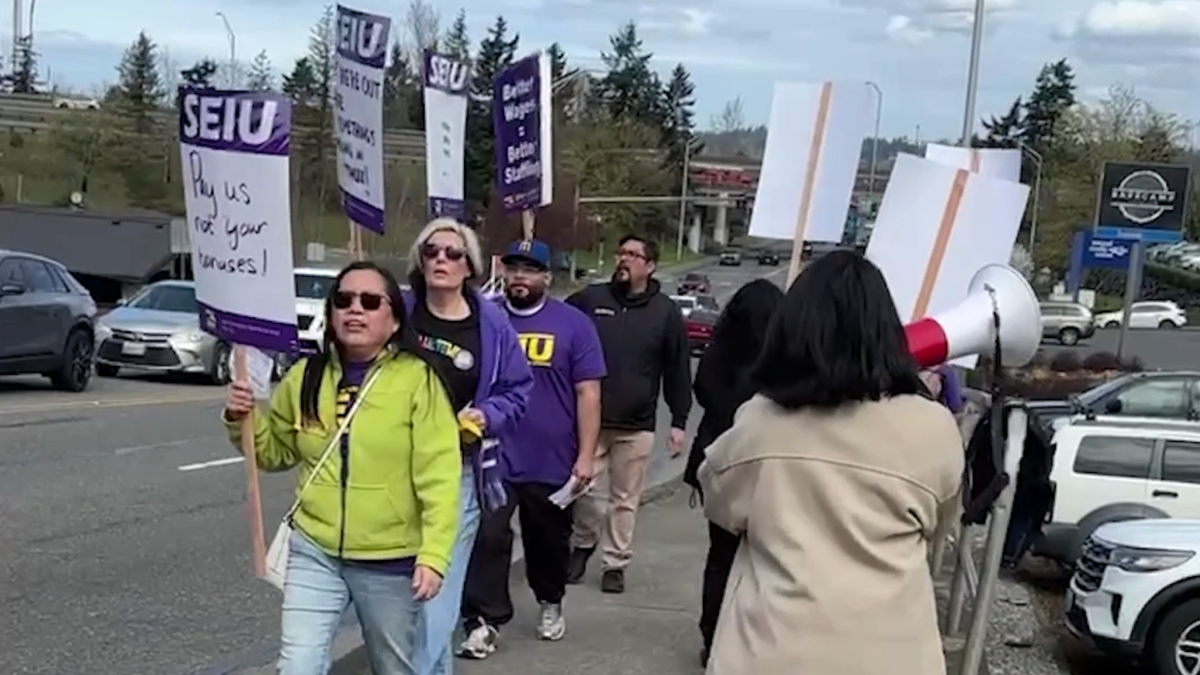 Group of protesters marching on a street, holding SEIU signs; a woman in a lime jacket leads with a sign. Meets a megaphone on the right.