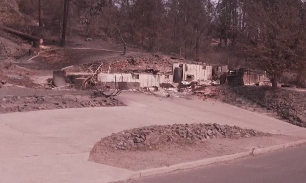 Residential street with collapsed, debris-filled foundations and damaged walls from a recent disaster; scattered rubble along the road edge.