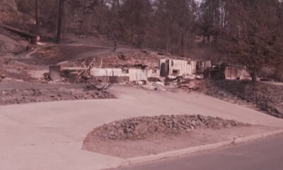 Residential street with collapsed, debris-filled foundations and damaged walls from a recent disaster; scattered rubble along the road edge.
