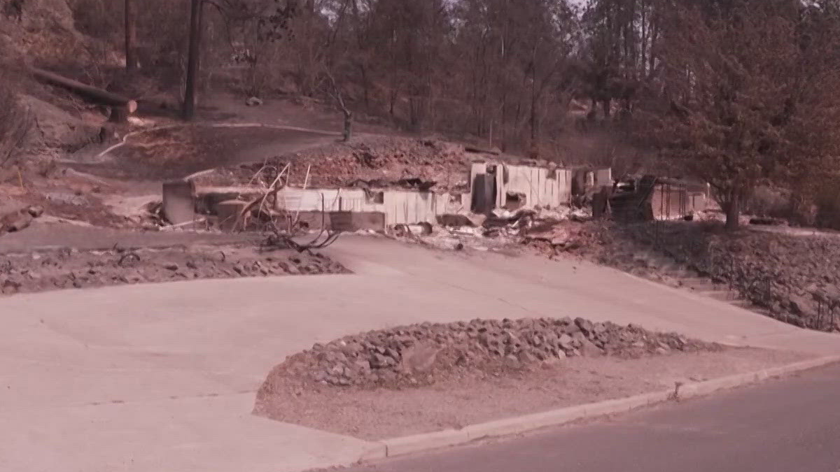 Residential street with collapsed, debris-filled foundations and damaged walls from a recent disaster; scattered rubble along the road edge.