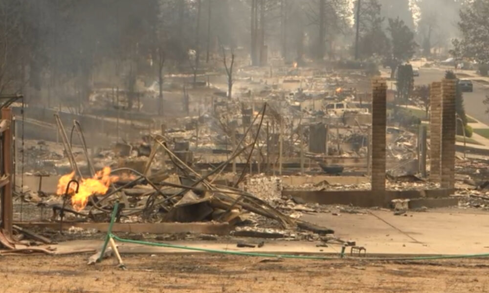 Charred remains of a house with flames still burning amid scattered debris in a suburban street scene.