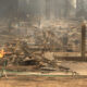 Charred remains of a house with flames still burning amid scattered debris in a suburban street scene.