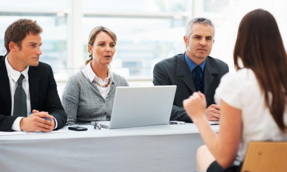 Panel of three professionals in business attire interviewing a candidate; a laptop and notes on the table.