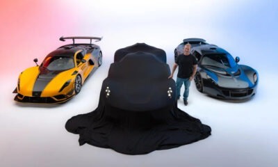 A black car is draped in a cover between a yellow and a blue sports car in a bright studio.