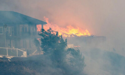 Fire and smoke engulf a multi‑story building on the left as flames consume structures to the right, with a fence in the foreground.