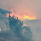 Fire and smoke engulf a multi‑story building on the left as flames consume structures to the right, with a fence in the foreground.