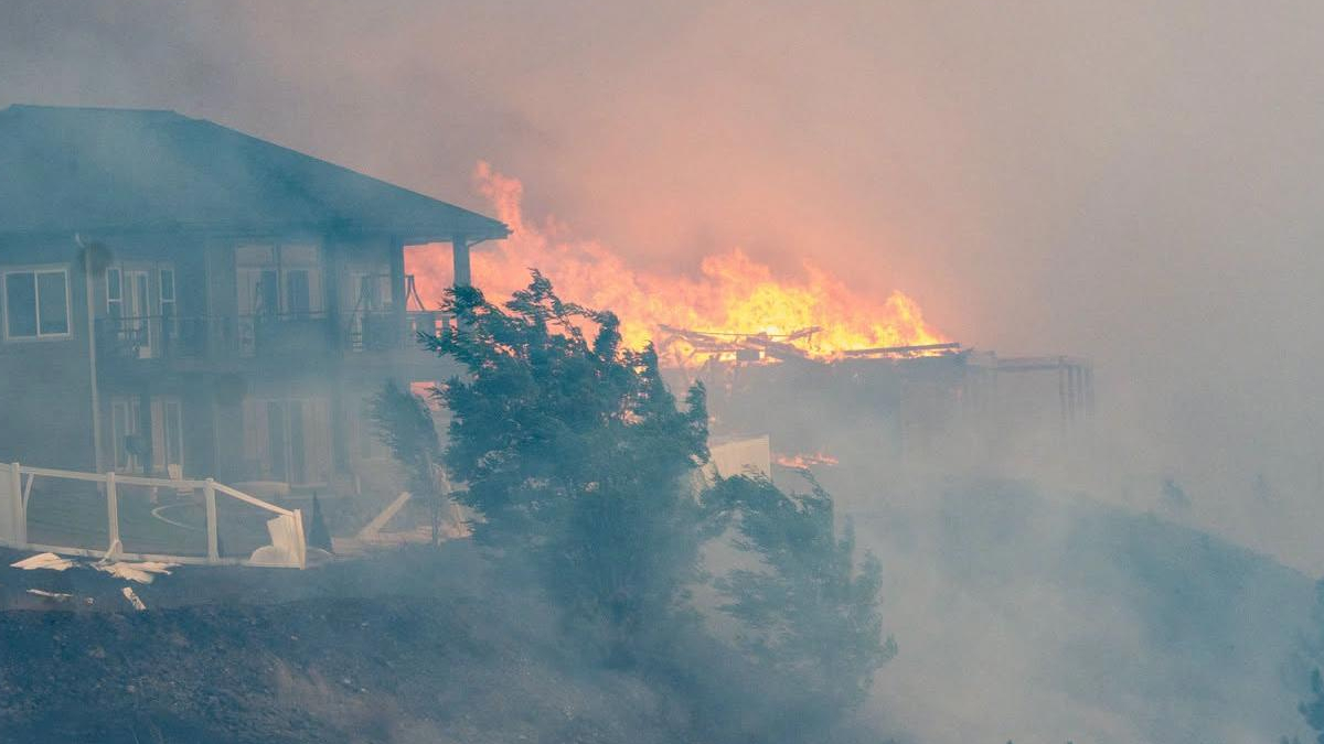 Fire and smoke engulf a multi‑story building on the left as flames consume structures to the right, with a fence in the foreground.