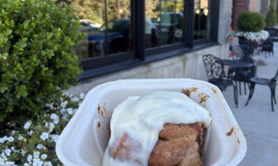 To-go container holding a glazed cinnamon roll outdoors near a sidewalk cafe with white flowers and black patio furniture in the background.