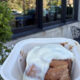 To-go container holding a glazed cinnamon roll outdoors near a sidewalk cafe with white flowers and black patio furniture in the background.