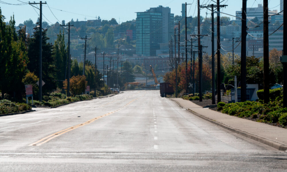 Wide empty street in a city neighborhood, power poles line both sides with trees and distant buildings on the skyline.