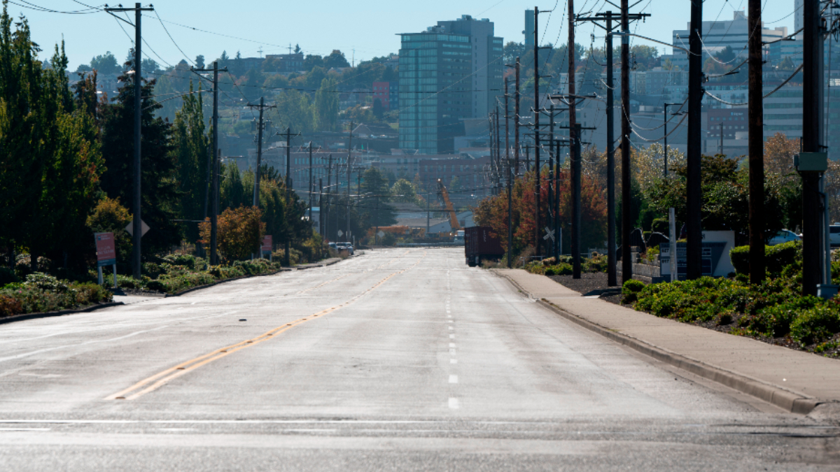 Wide empty street in a city neighborhood, power poles line both sides with trees and distant buildings on the skyline.