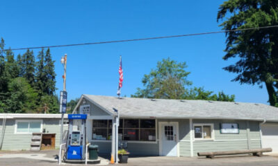 Small gray roadside gas station/general store with a blue gas pump, American flag, and a long wooden bench outside on a sunny day