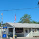 Small gray roadside gas station/general store with a blue gas pump, American flag, and a long wooden bench outside on a sunny day