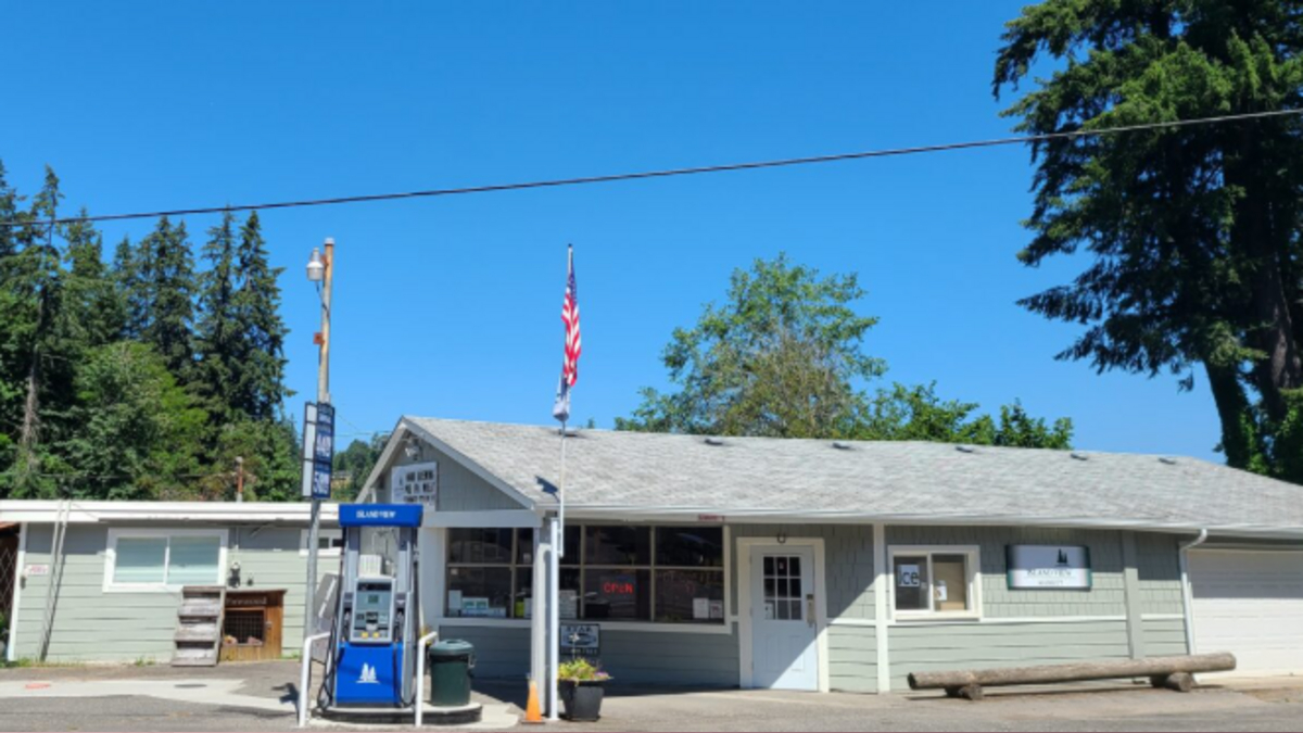 Small gray roadside gas station/general store with a blue gas pump, American flag, and a long wooden bench outside on a sunny day