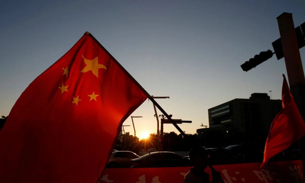 Red Chinese flag with yellow stars waving in the foreground as a sunset silhouettes a city street and cars behind it.