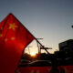 Red Chinese flag with yellow stars waving in the foreground as a sunset silhouettes a city street and cars behind it.