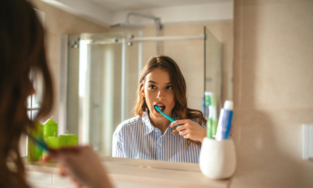 Woman brushing her teeth at bathroom sink, seen in the mirror, with a blue toothbrush.