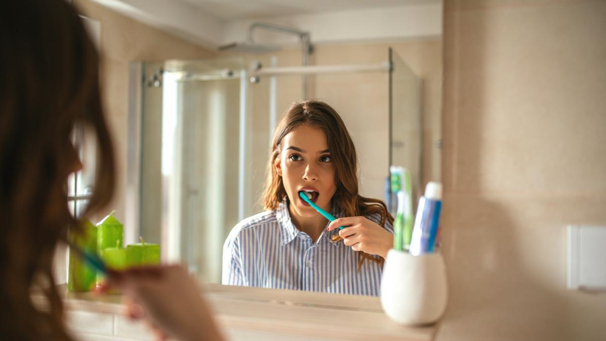 Woman brushing her teeth at bathroom sink, seen in the mirror, with a blue toothbrush.