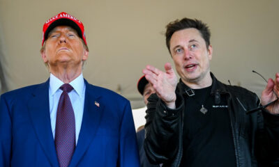 Two men stand under a tent; one in a blue suit and red MAGA hat looks upward, the other in a black jacket gestures while holding glasses.