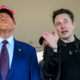 Two men stand under a tent; one in a blue suit and red MAGA hat looks upward, the other in a black jacket gestures while holding glasses.