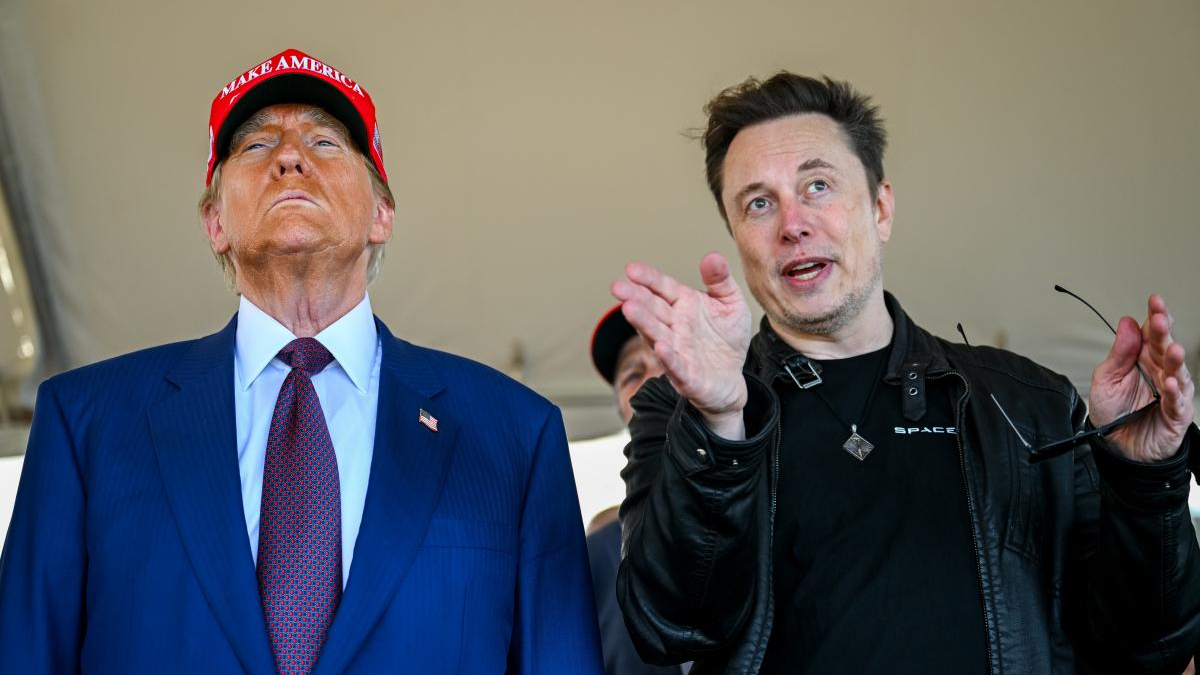 Two men stand under a tent; one in a blue suit and red MAGA hat looks upward, the other in a black jacket gestures while holding glasses.