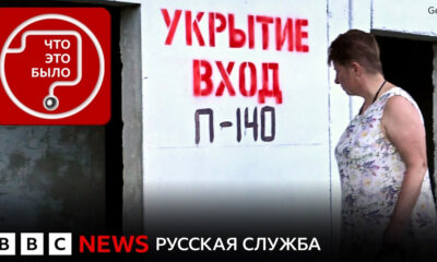 A woman in a floral sleeveless dress stands at a doorway as red Cyrillic signage reads 'УКРЫТИЕ ВХОД П-140' on a white wall, BBC News watermark visible.