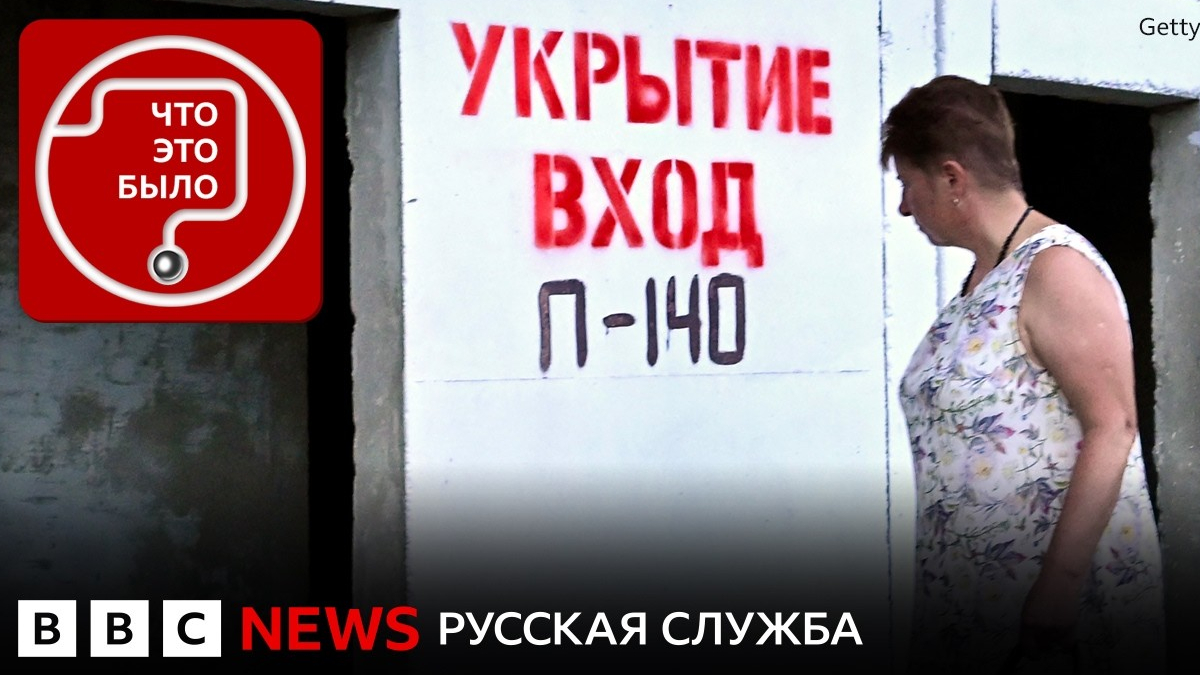 A woman in a floral sleeveless dress stands at a doorway as red Cyrillic signage reads 'УКРЫТИЕ ВХОД П-140' on a white wall, BBC News watermark visible.