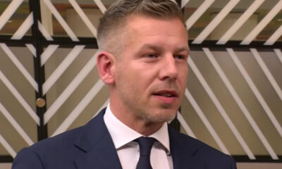 Man in a navy suit and tie speaking in a studio with a striped geometric backdrop behind him.