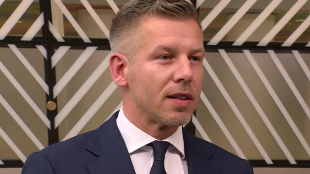 Man in a navy suit and tie speaking in a studio with a striped geometric backdrop behind him.