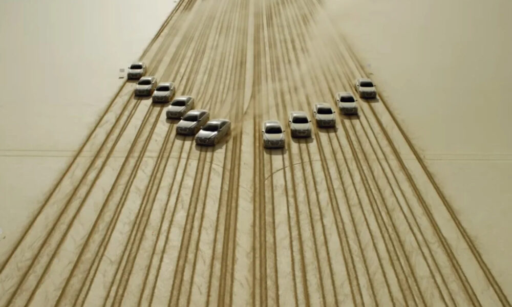 Aerial view of cars arranged in a curved V pattern on dusty desert sand, with visible tire tracks across the surface.