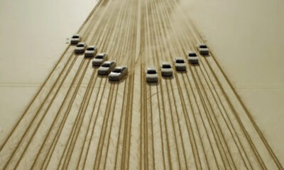 Aerial view of cars arranged in a curved V pattern on dusty desert sand, with visible tire tracks across the surface.