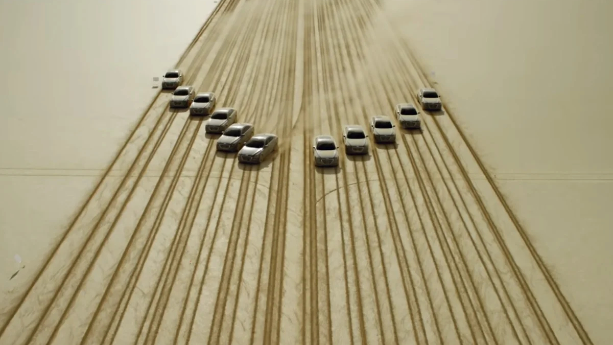 Aerial view of cars arranged in a curved V pattern on dusty desert sand, with visible tire tracks across the surface.