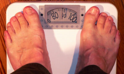 Top-down view of a person's bare feet standing on a white analog bathroom scale, showing a weight around 95. 00 pounds/units marked on the dial are visible.