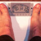 Top-down view of a person's bare feet standing on a white analog bathroom scale, showing a weight around 95. 00 pounds/units marked on the dial are visible.