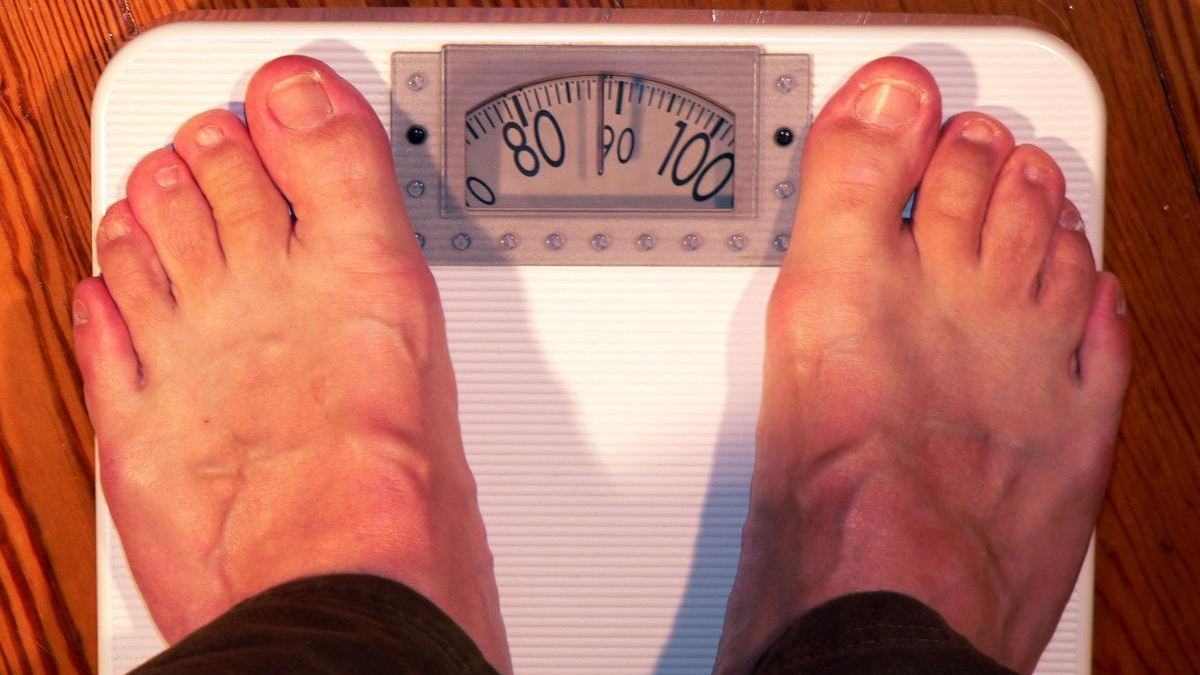 Top-down view of a person's bare feet standing on a white analog bathroom scale, showing a weight around 95. 00 pounds/units marked on the dial are visible.