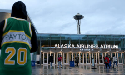 Person in a green jersey with 'PAYTON' and the number 20 stands at the left, facing Alaska Airlines Atrium entrance with the Space Needle in the distance on a cloudy day.