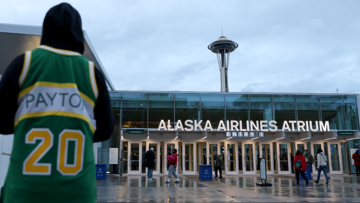 Person in a green jersey with 'PAYTON' and the number 20 stands at the left, facing Alaska Airlines Atrium entrance with the Space Needle in the distance on a cloudy day.