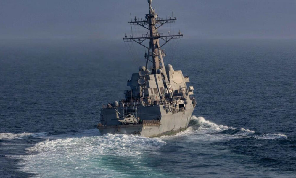 Grey naval warship cutting through blue ocean, tall radar mast and deck sensors visible at the fore. (informative)