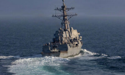 Grey naval warship cutting through blue ocean, tall radar mast and deck sensors visible at the fore. (informative)
