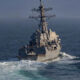 Grey naval warship cutting through blue ocean, tall radar mast and deck sensors visible at the fore. (informative)