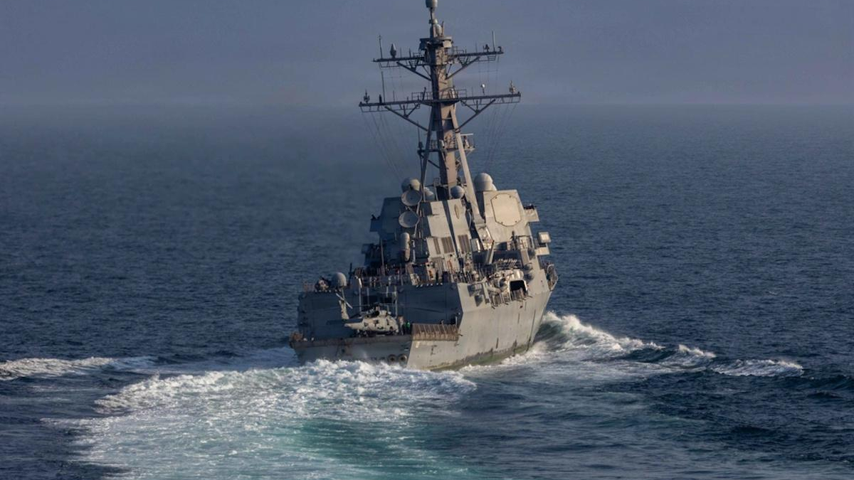 Grey naval warship cutting through blue ocean, tall radar mast and deck sensors visible at the fore. (informative)