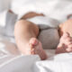 Close-up of a baby's feet resting on a white bedsheet, wearing a striped diaper piece visible above the blanket.