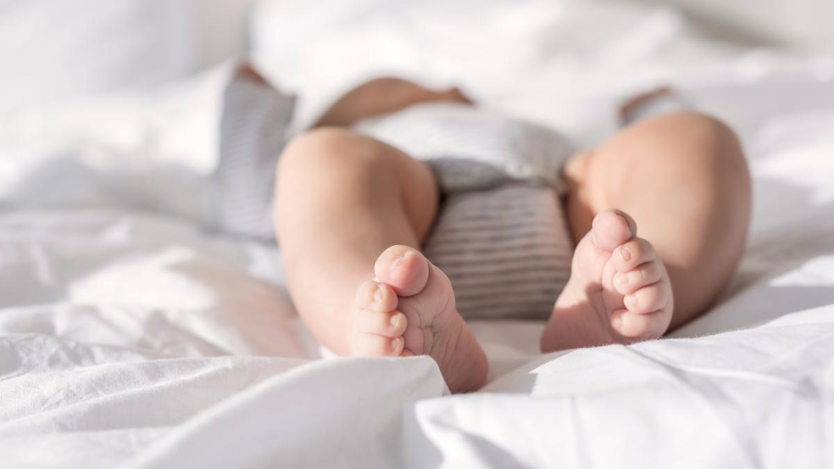 Close-up of a baby's feet resting on a white bedsheet, wearing a striped diaper piece visible above the blanket.