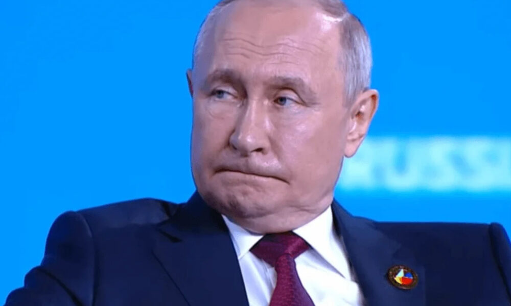 Man in a dark suit and burgundy tie seated on a stage against a bright blue background.