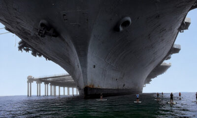 Low-angle view of a colossal ship hull towering above the water with several paddleboarders in the foreground.