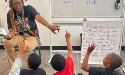 Teacher reading aloud to a group of students who raise their hands in a classroom during a read-aloud lesson with a reading poster nearby.