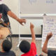Teacher reading aloud to a group of students who raise their hands in a classroom during a read-aloud lesson with a reading poster nearby.